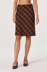 A woman is wearing a knee-length skirt with a brown plaid pattern, featuring intersecting yellow and dark brown lines. The skirt has a small bow detail at the waistband. She is also wearing brown heeled shoes with ankle straps.